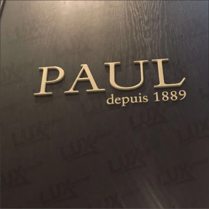 Объёмная вывеска PAUL depuis 1889 из светлых букв на тёмной стеновой панели с выраженной древесной текстурой.